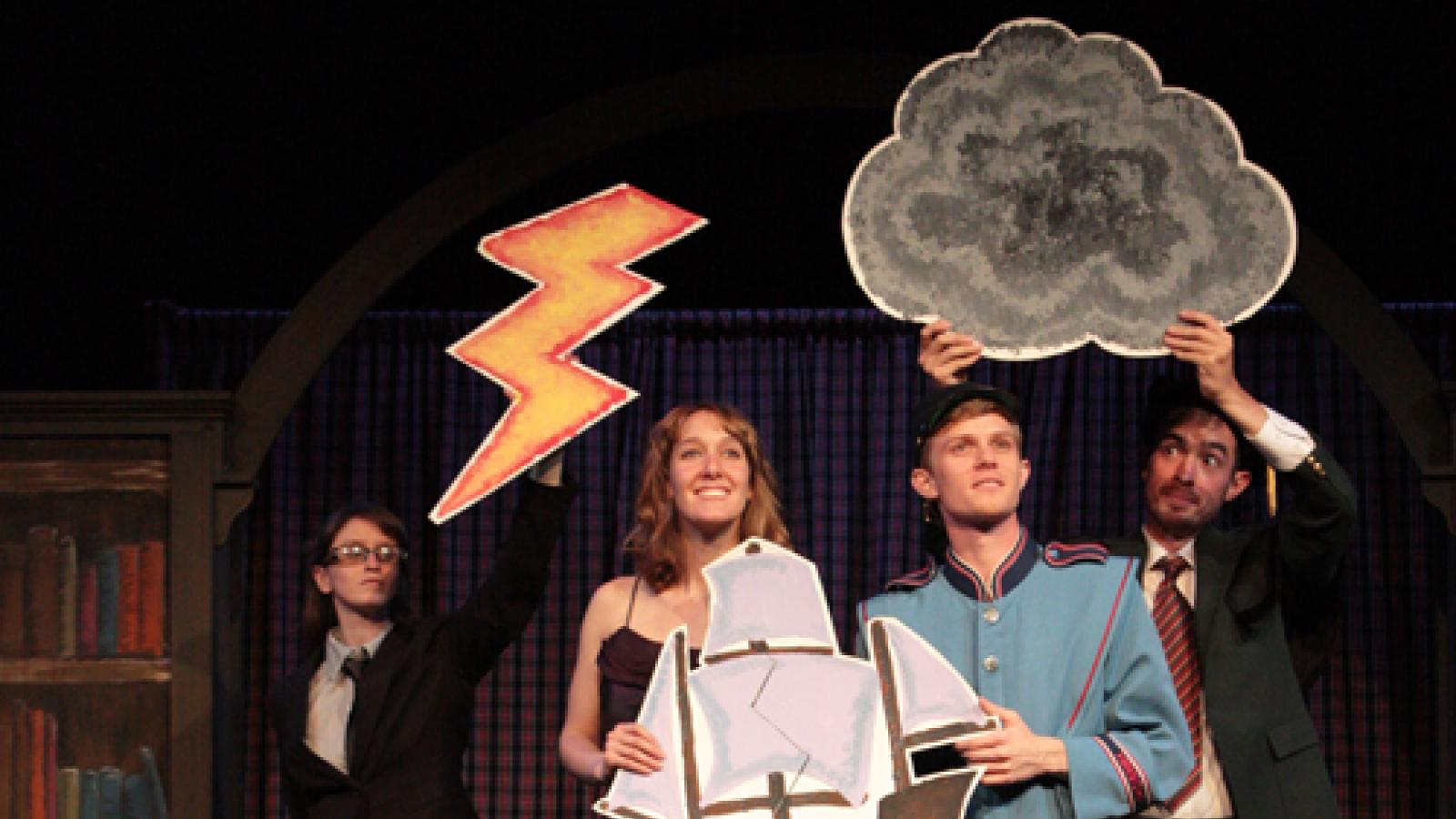 Six actors performing, holding cut outs of waves, a ship, clouds, and lightning