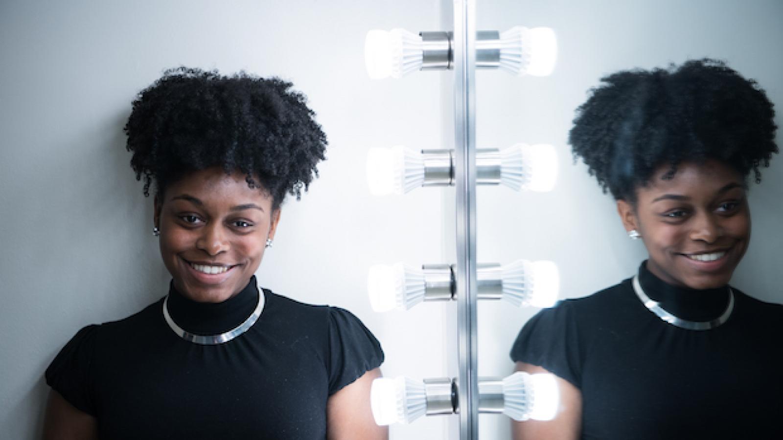 a young African American woman with an updo and a black top stands next to a dressing room mirror so her image is doubled