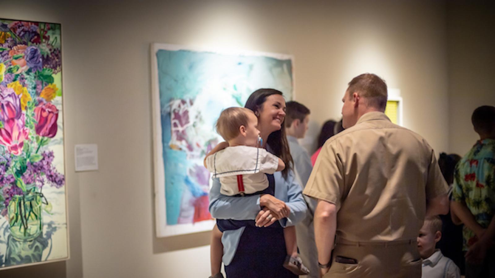 a man and a woman holding a child talk to each other against a background of colorful paintings