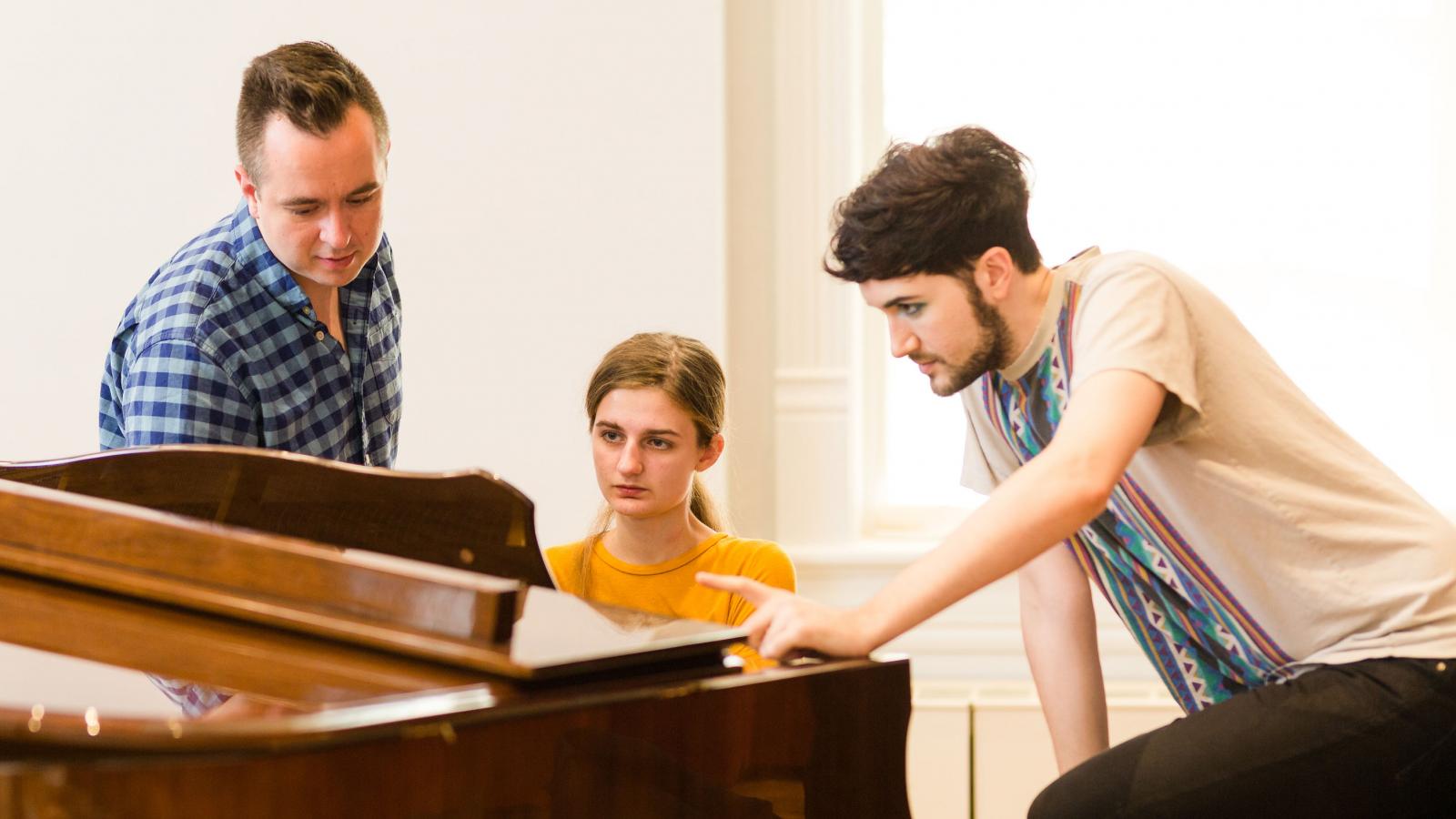 female student sits at piano with two men looking at sheet music with her