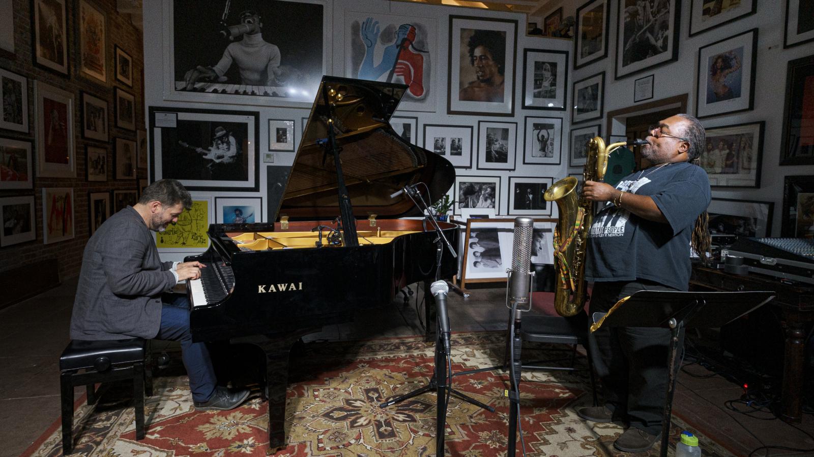 A man plays piano and another plays saxophone in a small art gallery