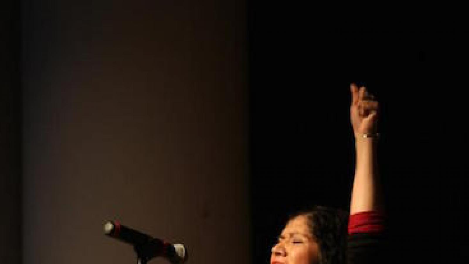 an indigenous American woman stands in front of a microphone one arm thrust in the air