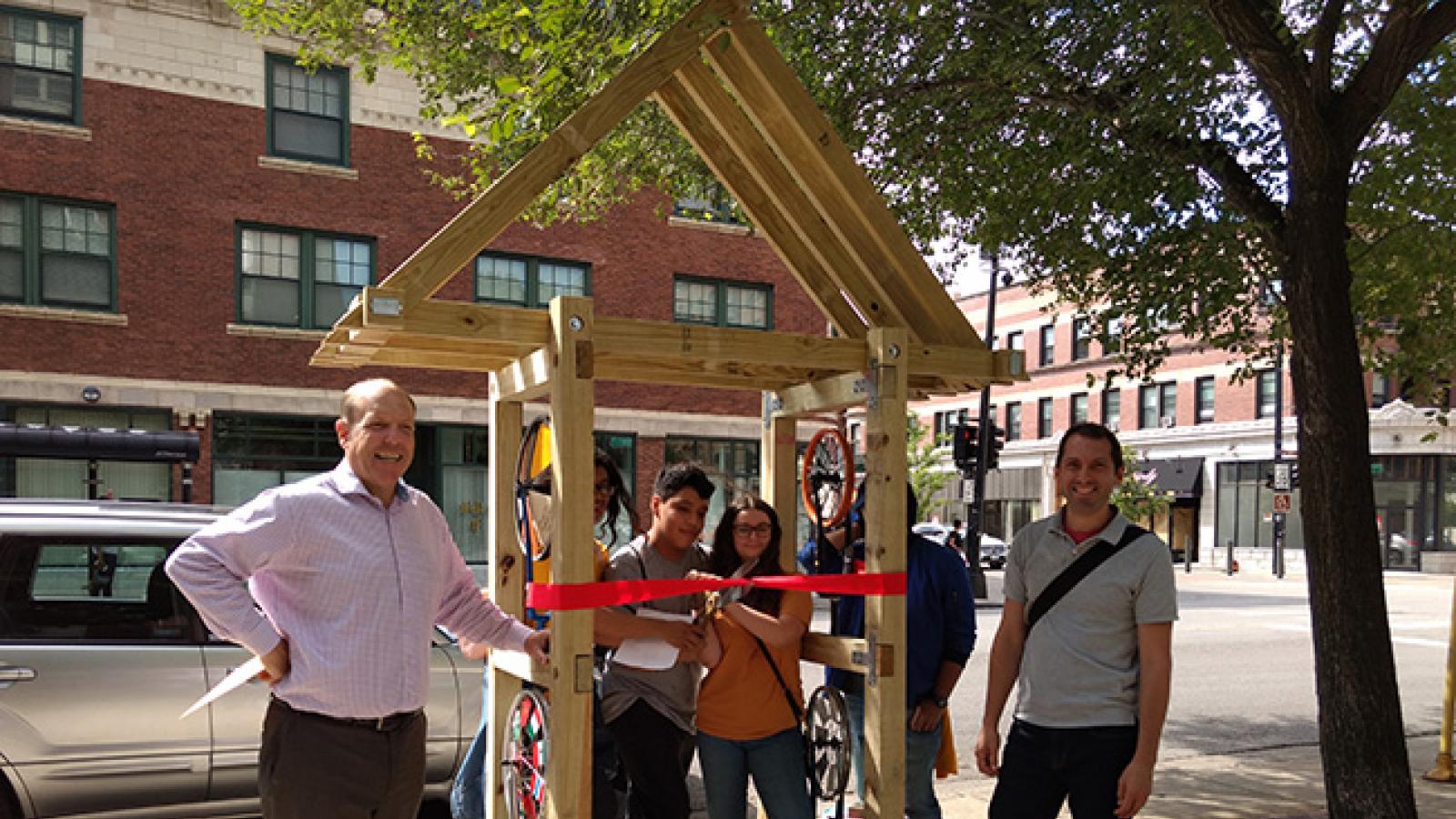 Two teens cut a ribbon on a newly installed arrow sculpture while two adult men stand smiling at the camera