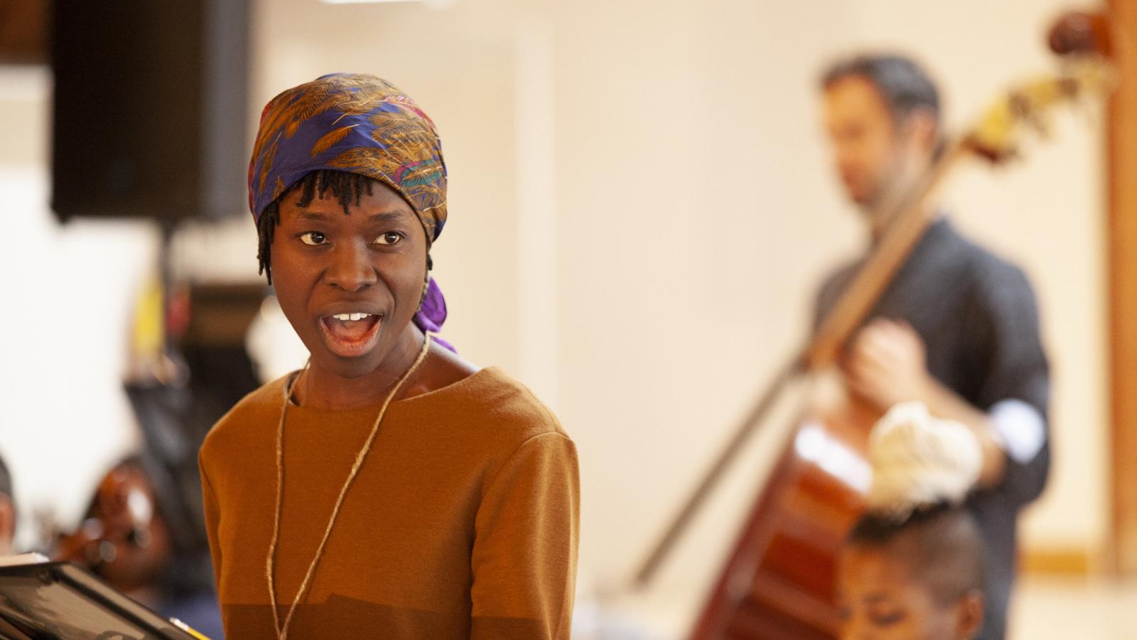 A woman rehearses singing in front of a music stand with an orchestra behind her