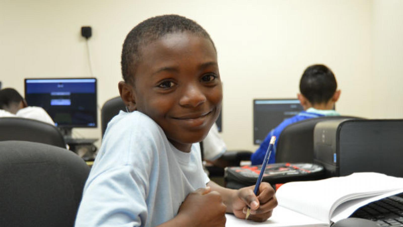 A child writing in a composition book and smiling at the camera