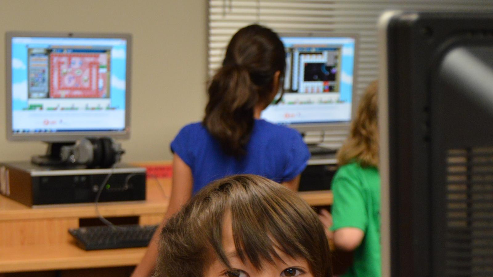 A young boy sits in the front of a computer with other students working on computers in the background.