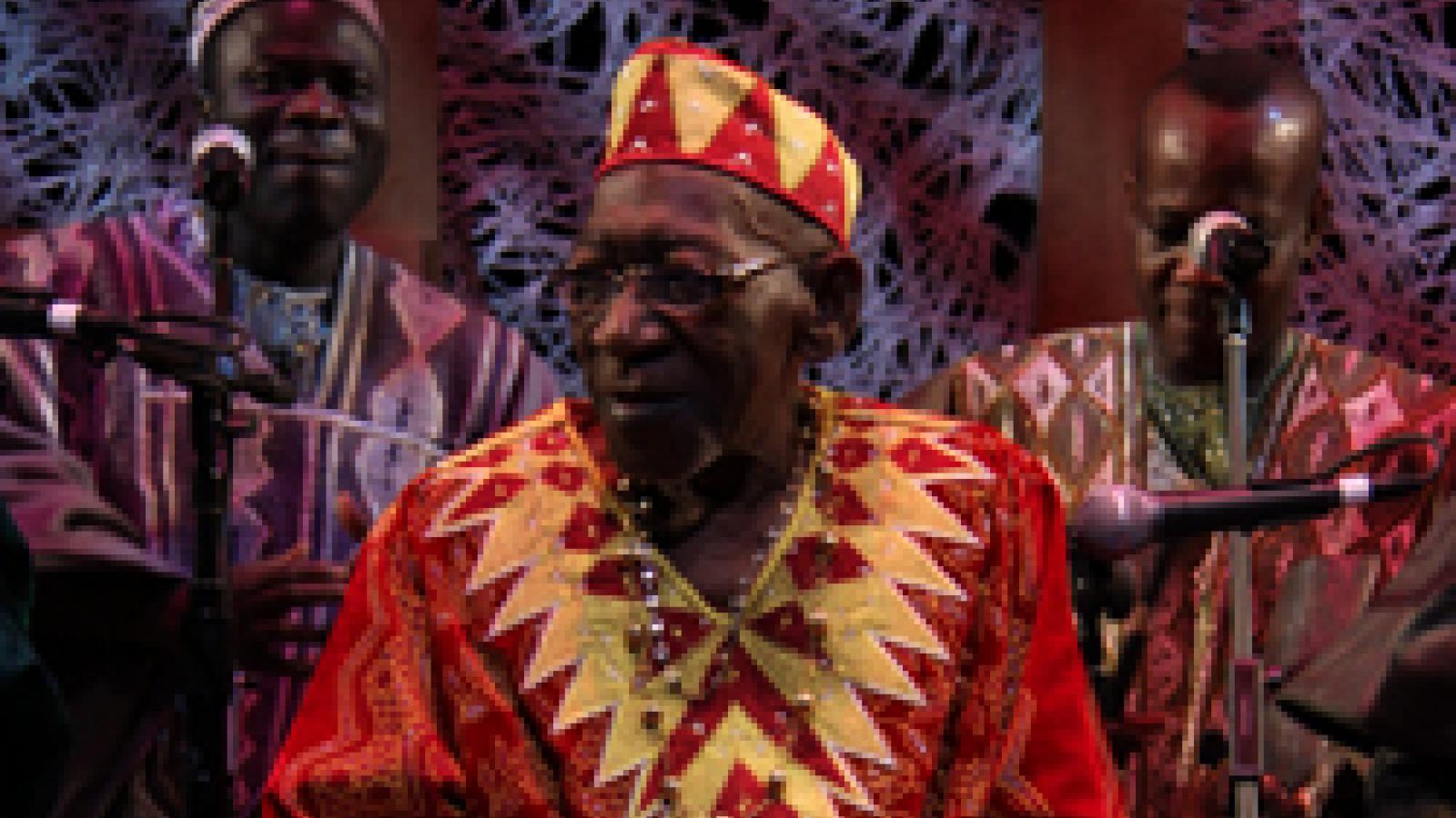 A man in a brightly colored red and yellow outfit plays a large drum with his hands.