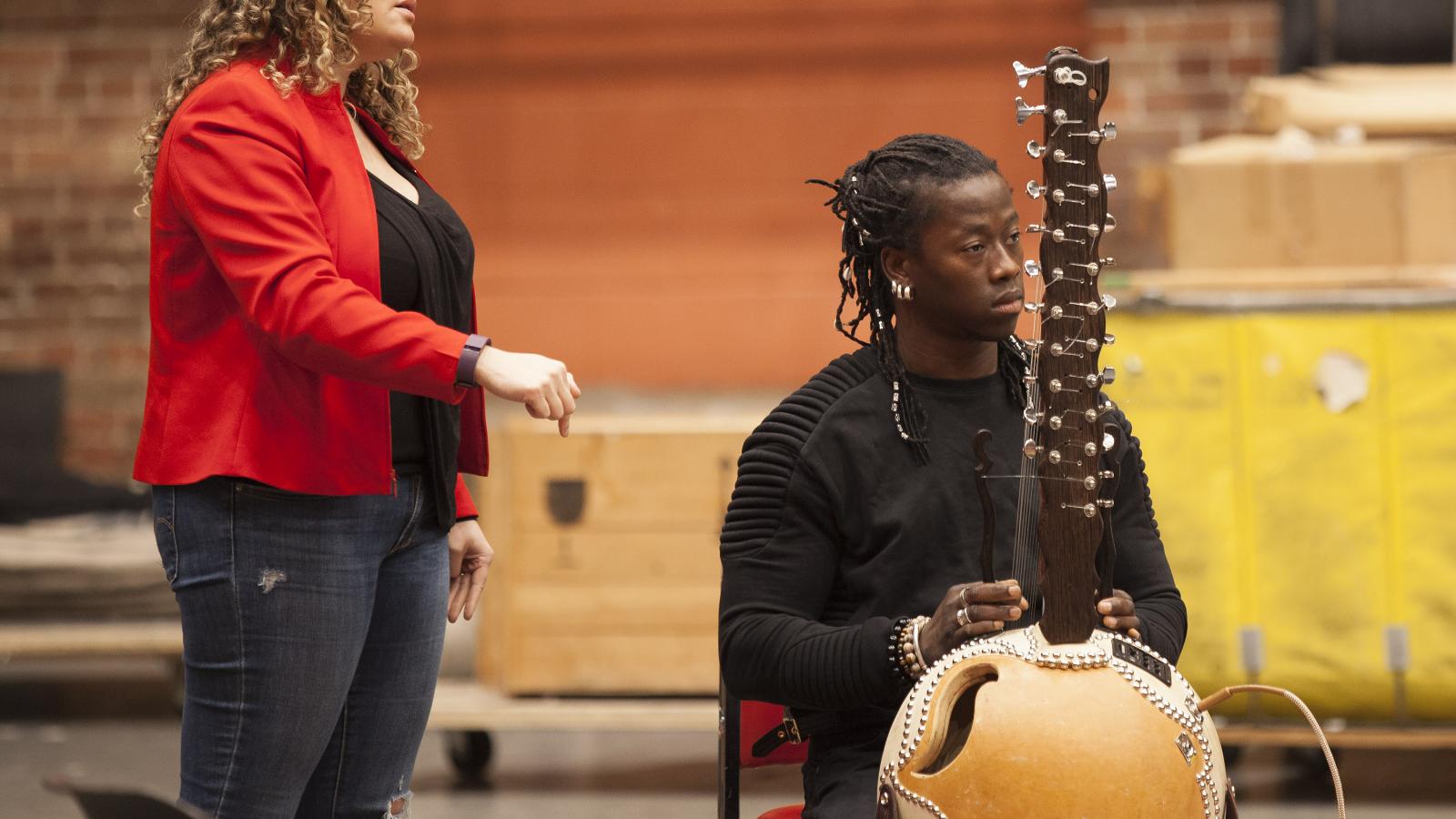 Woman standing next to a man playing a kora instrument.