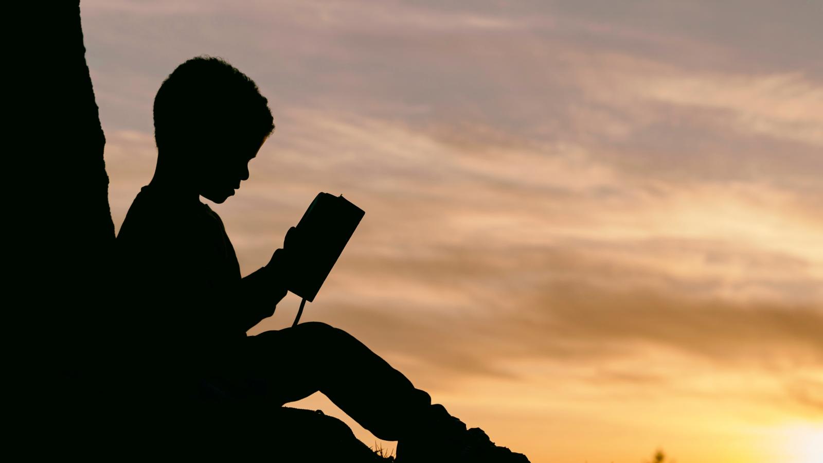 Silhouette of young boy reading a book against the sunset