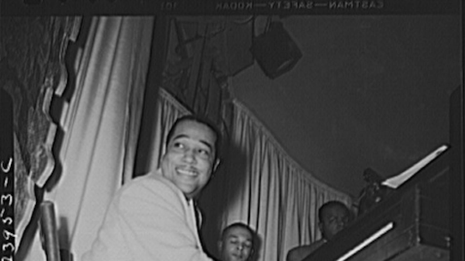 Duke Ellington seated at a piano smiling at someone offscreen as he plays