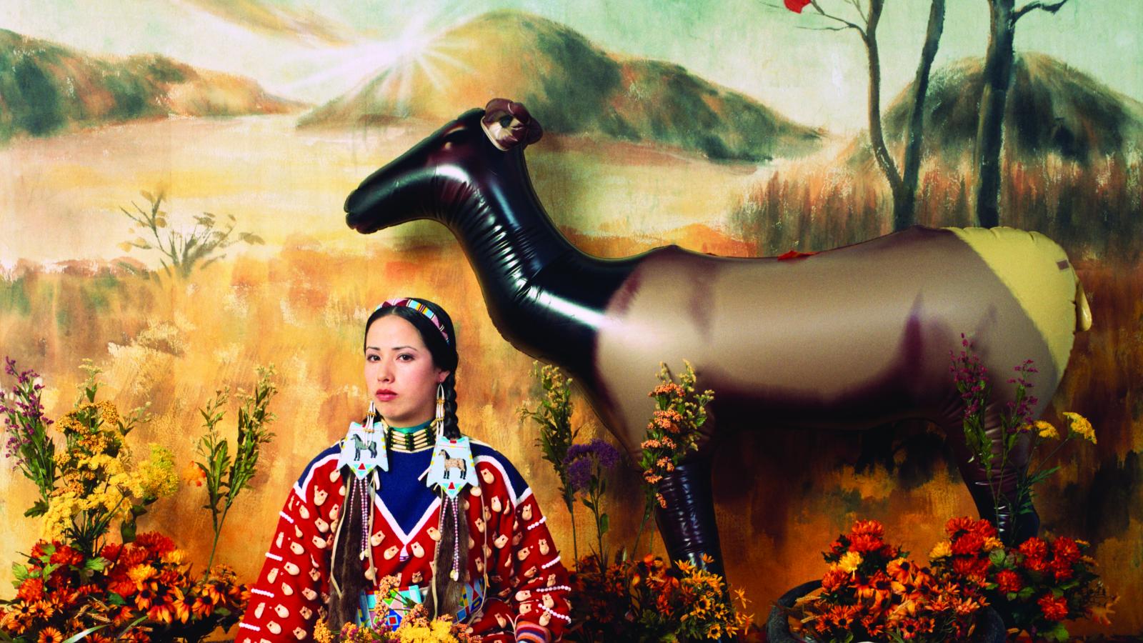 Woman in Native outfit sitting in front of a plains background  with a cow skull next to her and an inflatable elk behind her. 