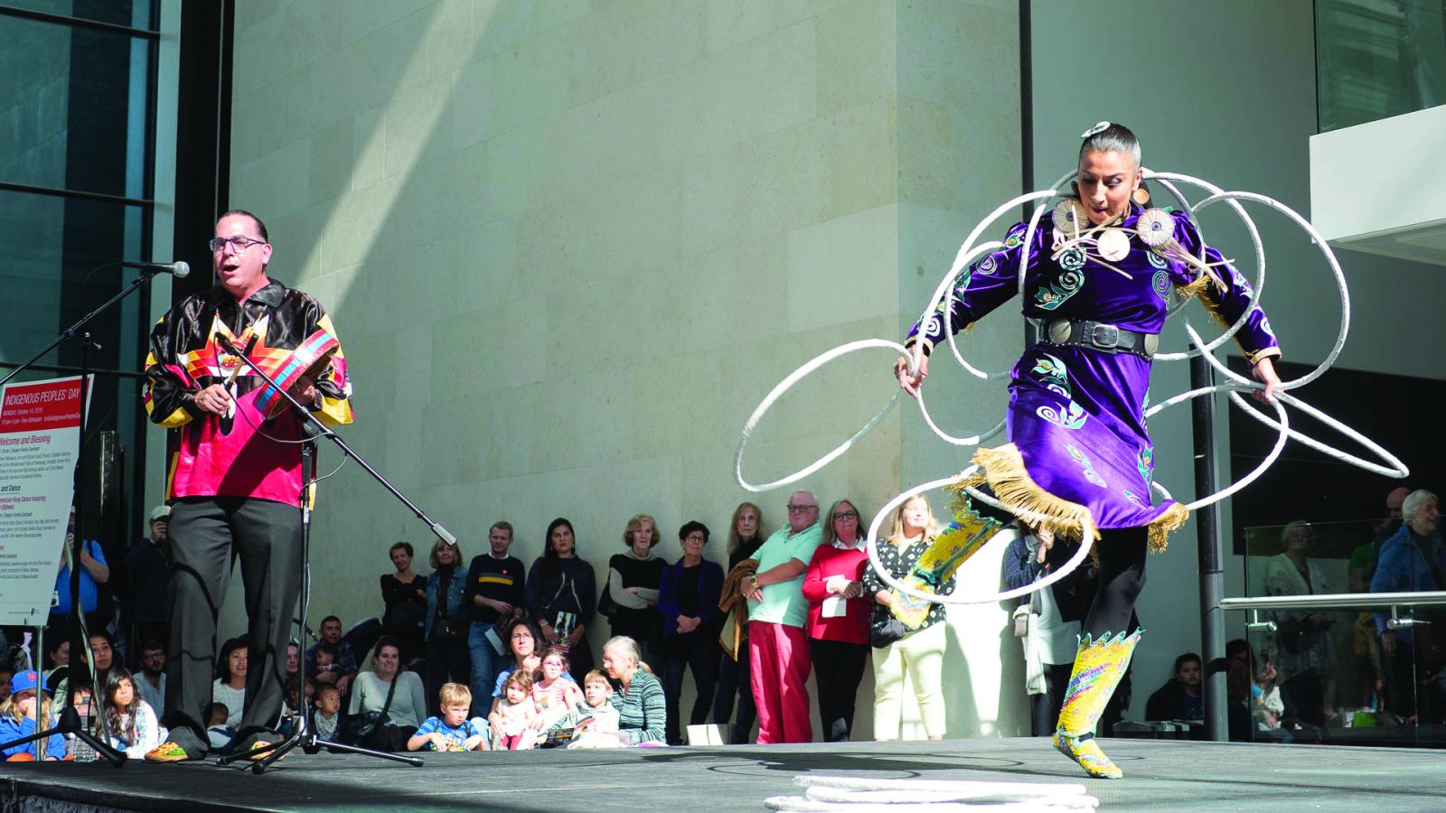 Woman in purple outfit dancing with hoops on stage while man to her right bangs and drum and sings. 