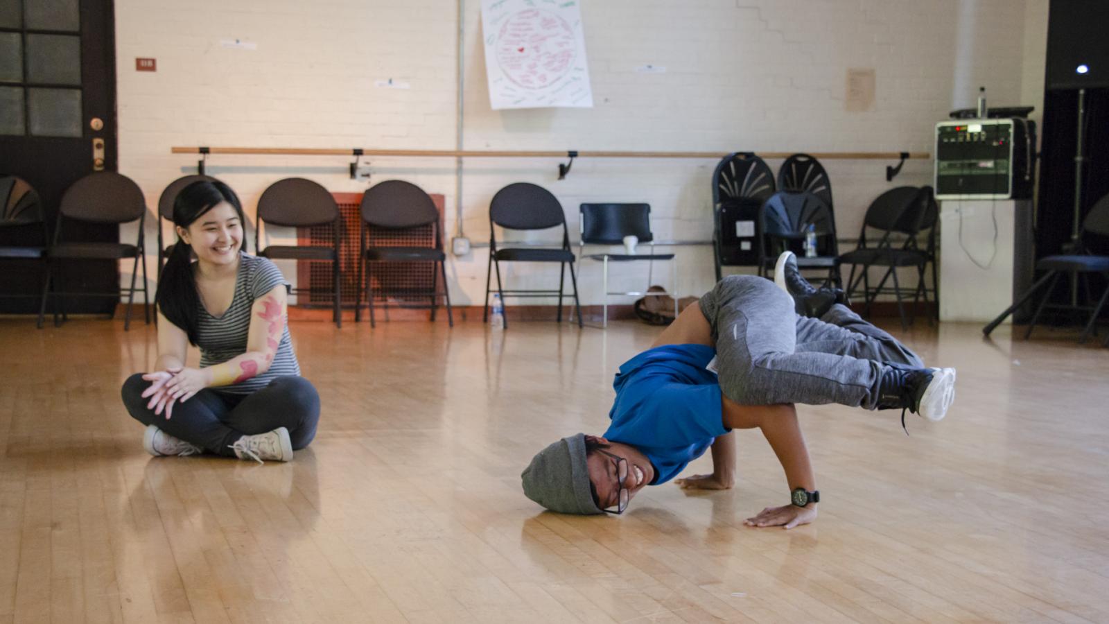 Student wearing hat dancing while a girl sitting cross-legged nearby watches.