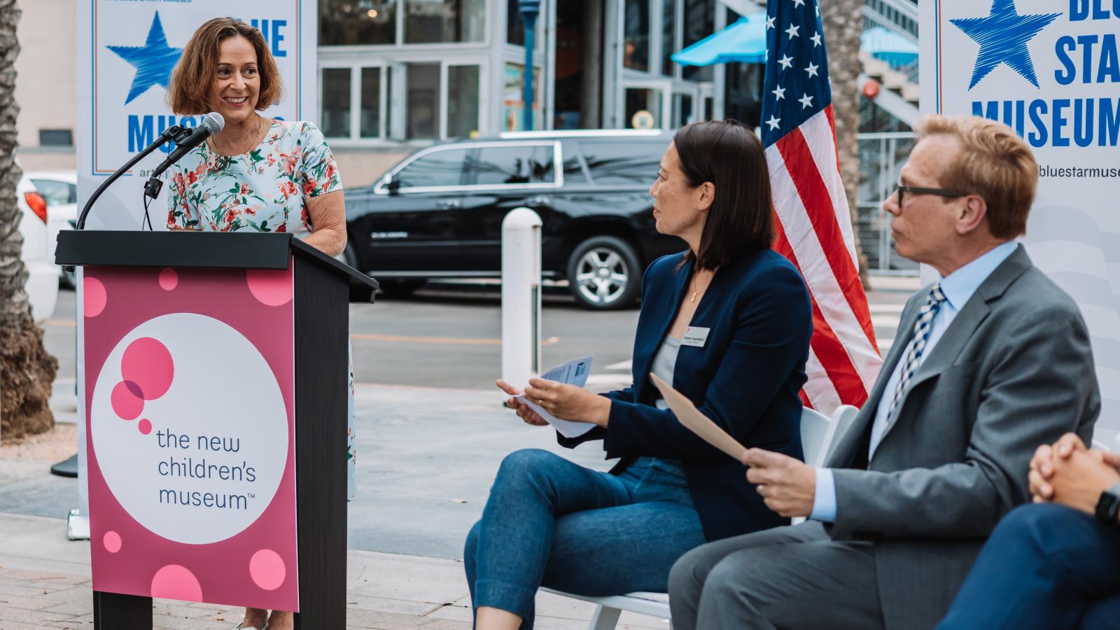 Woman to the left in front of a lectern with two people sitting watching her.