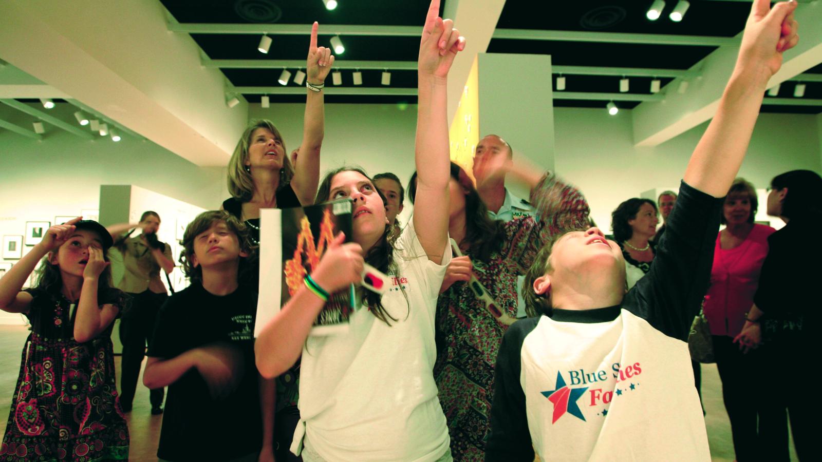 A group of children in a museum, all pointing and looking upward. 