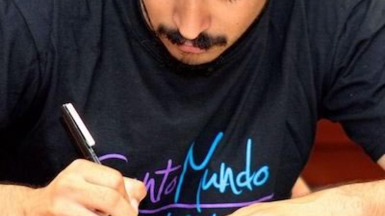 Headshot of a young Latino man wearing a hat and a Canto Mundo t-shirt bent over and writing on a page