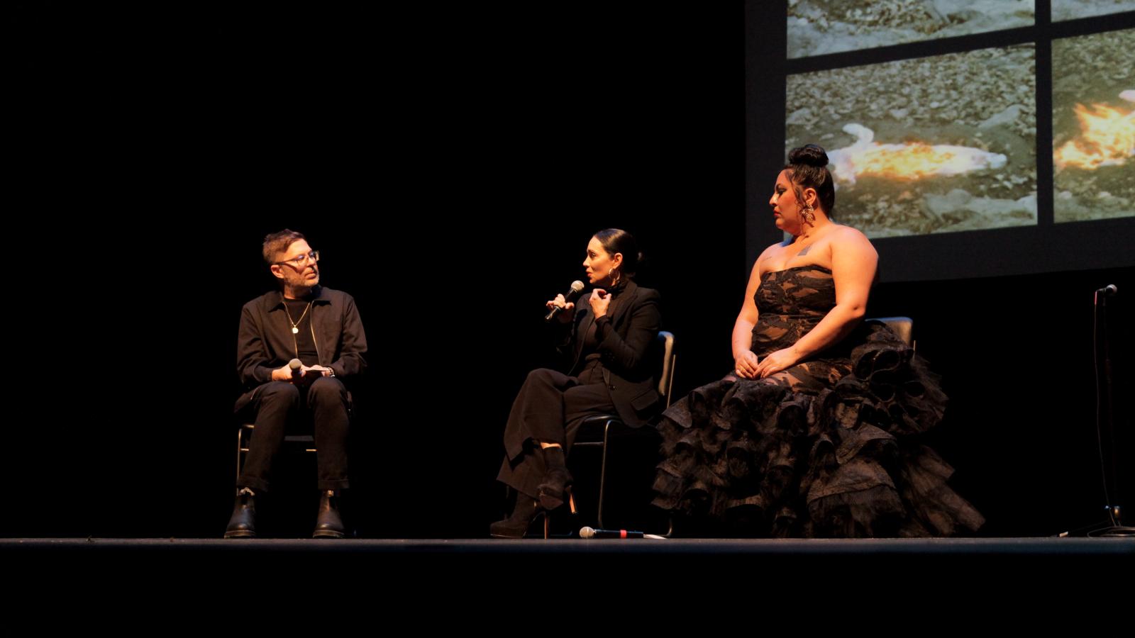 A man at left interviewing two woman dressed in black, one in a frilly dress on stage. 