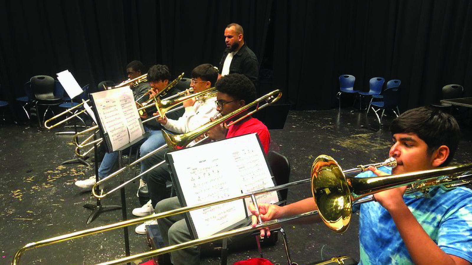 High school students playing trombone with musical stands near them, and man with shaved head and beard behind them instructing.