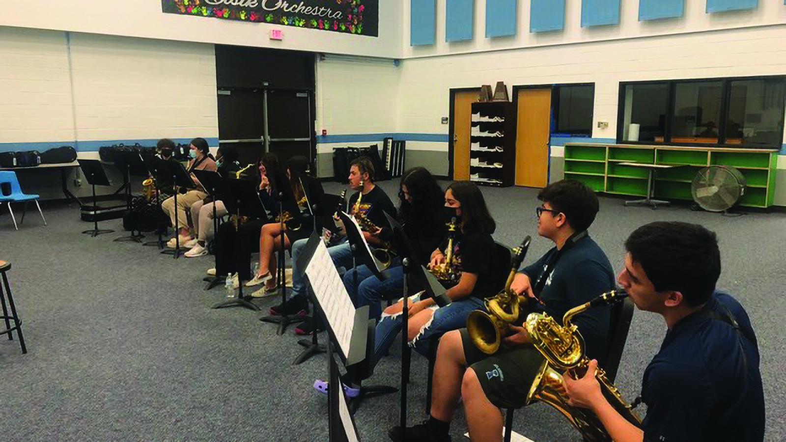 In a classroom, a line of high school students with musical stands in front of them playing saxophone.