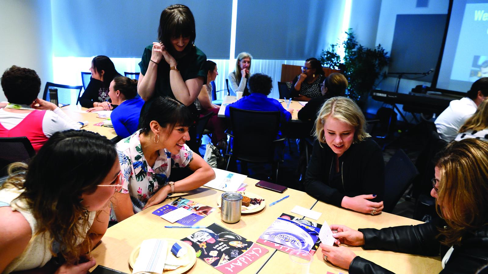 Four women sit around a table working on an art project while a woman stands behind them looking on.