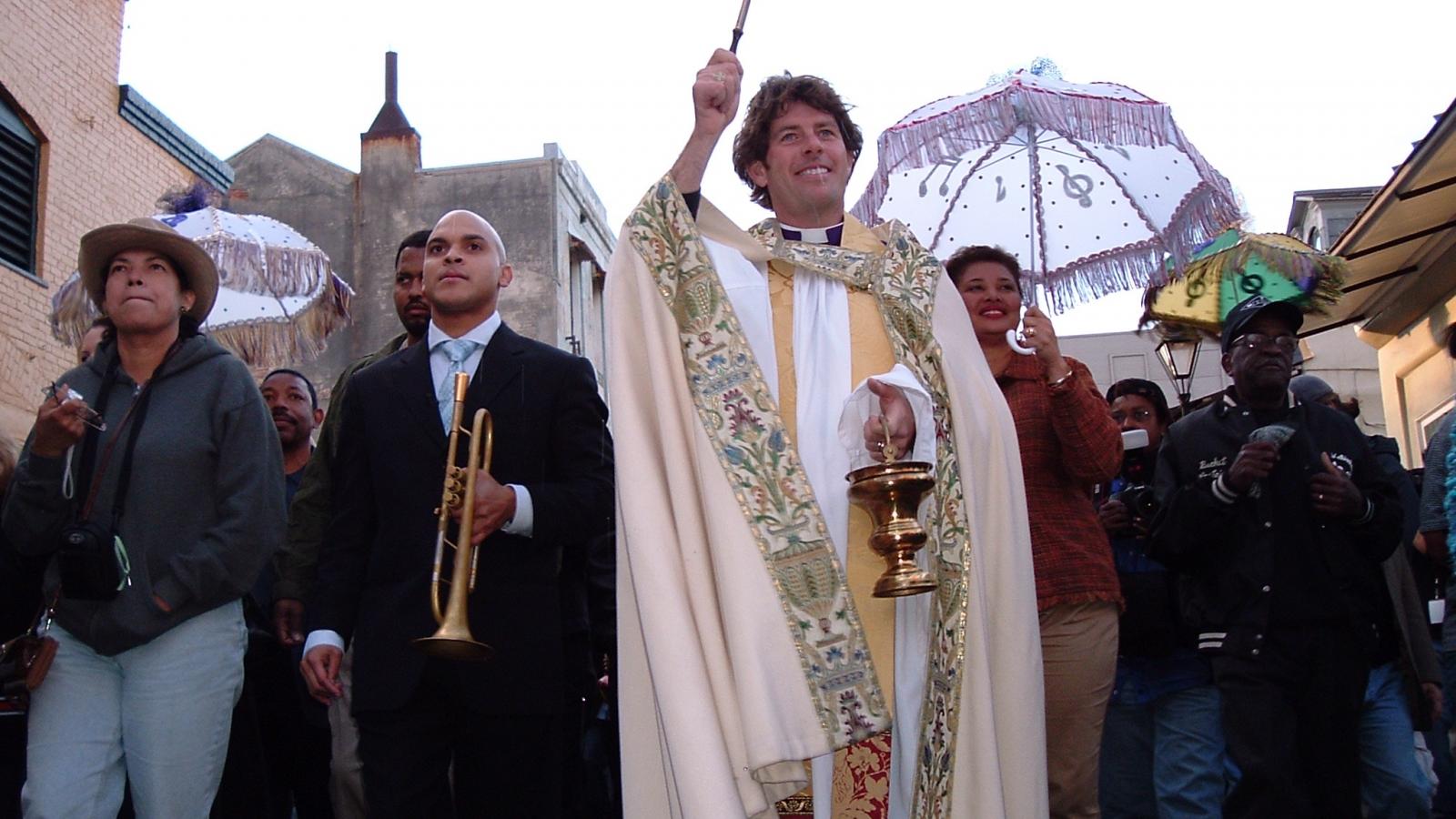 Man with trumpet next to priest blessing crowd in parade down street. 