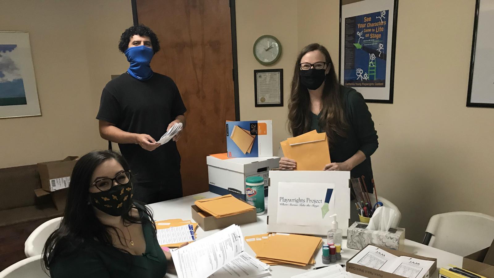 Playwrights Project Three people wearing masks look at the camera as they prepare large envelopes for mailing