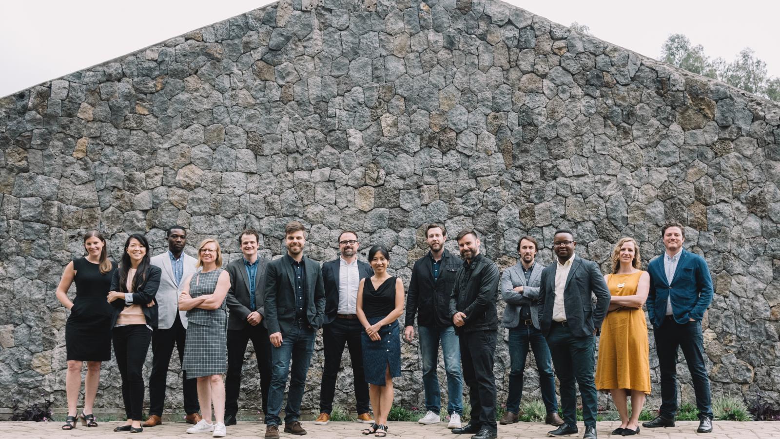 MASS Design Group A group of people stands smiling in front of a large stone wall
