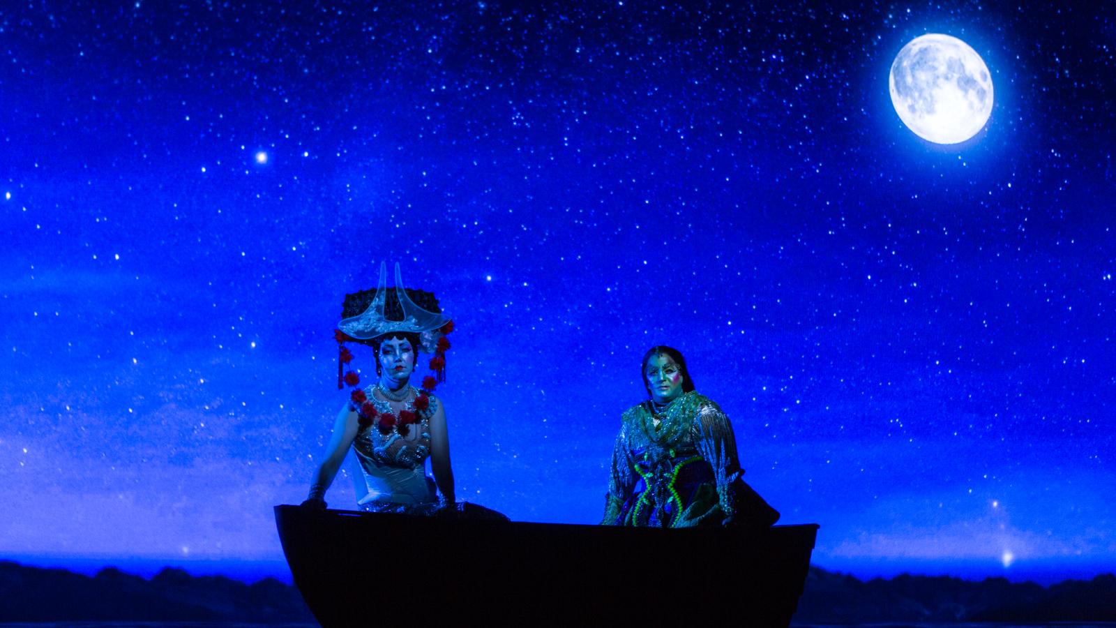 A stage production photo of two people ornately dressed in a boat with the moon behind them. 