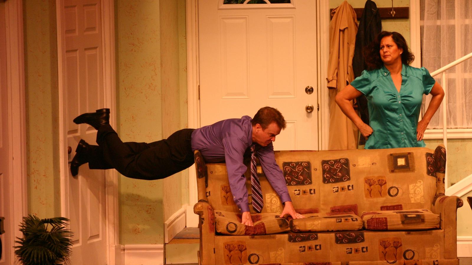 Man holding on to couch with feet on the wall while a woman with hands on hips watches. 