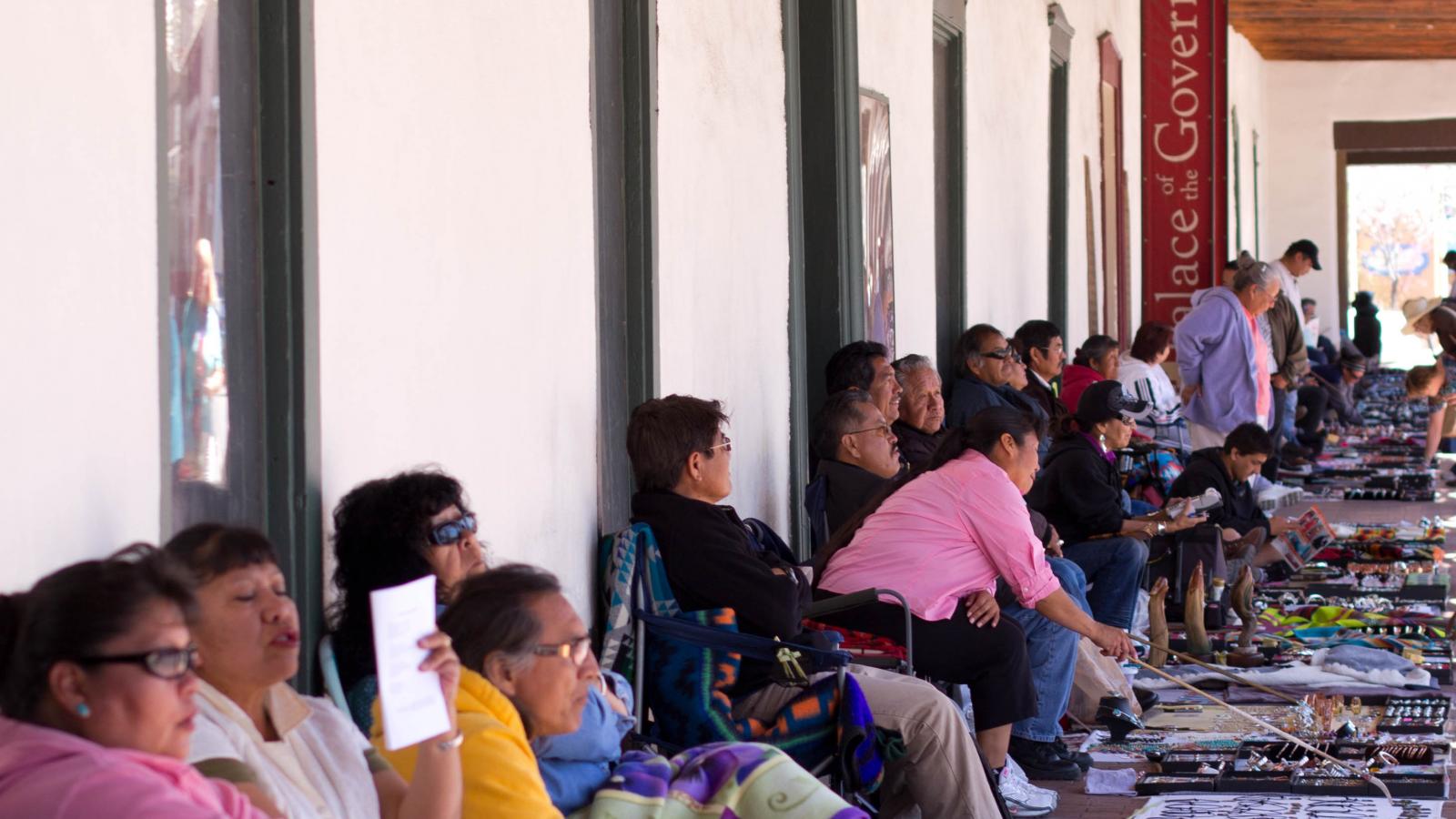 Native artists sit in chairs against a wall with their wares spread out on the sidewalk before them