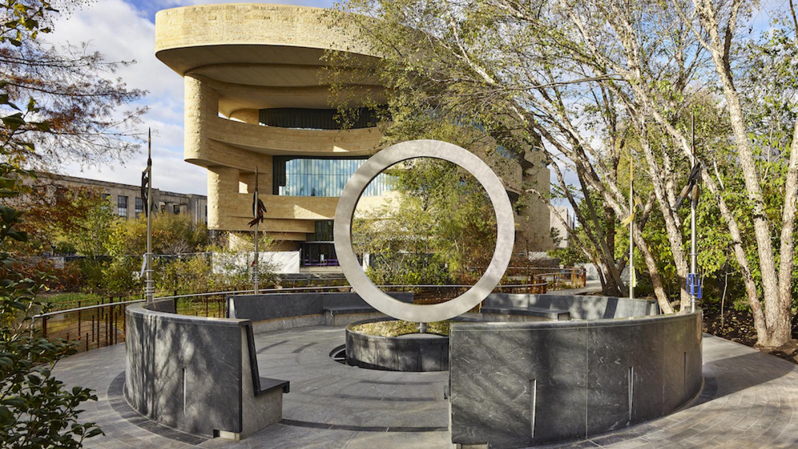 a view of the National Native American Veterans Memorial, which is a large circle on its side on top of a drum shape. The exterior of the National Museum of the American Indian is visible in the background