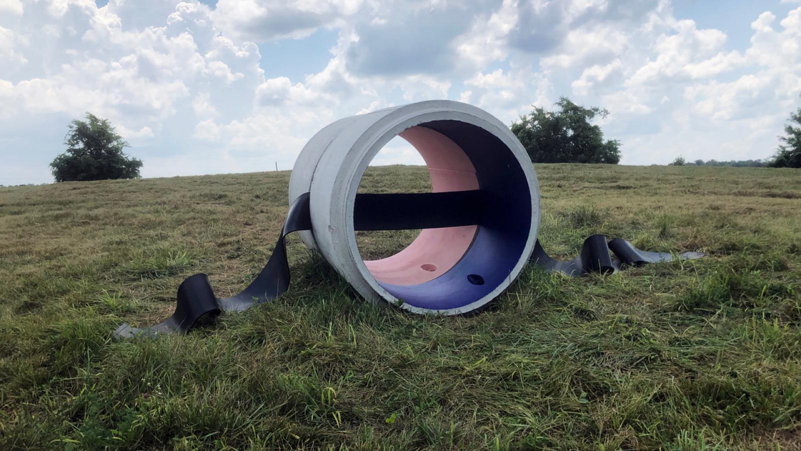 A barrel shaped sculpture with pink and purple interior sits in a grassy field