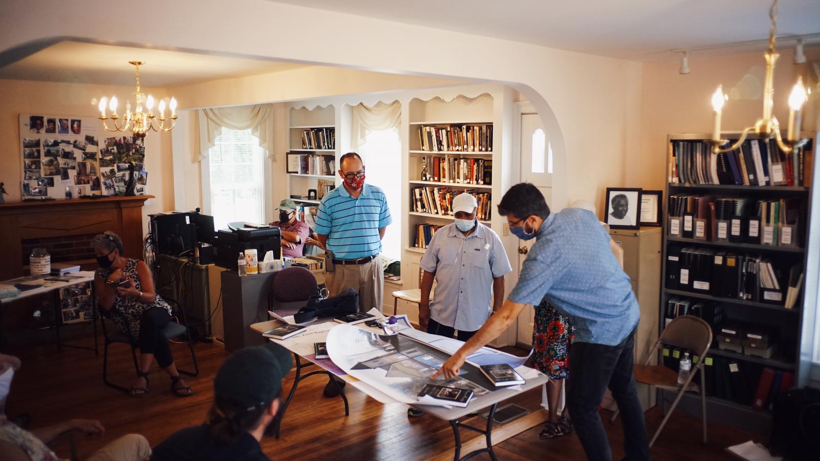 Three men wearing masks standing around a table with photos on it, one man pointing to one of the photos.