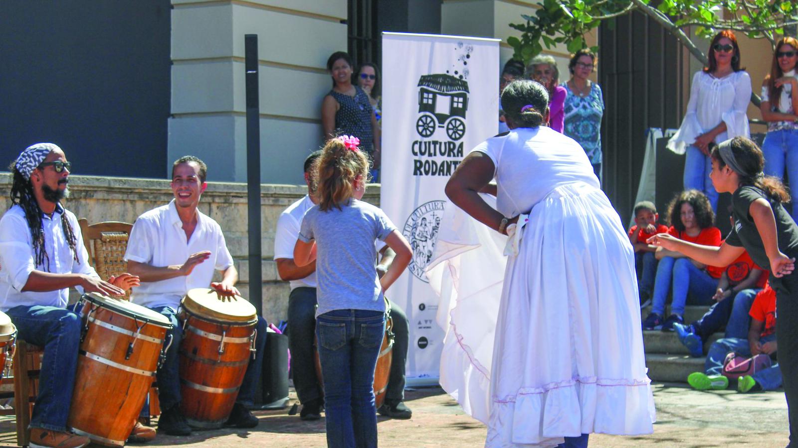 Woman in white dress teaching children to dance outdoors, with men playing drums in background.