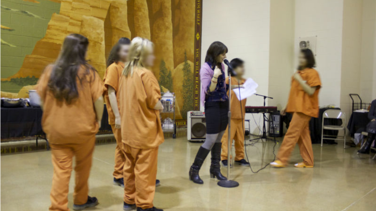 A woman with brown hair stands at the mic while girls in orange jumpsuits stand next to her. 