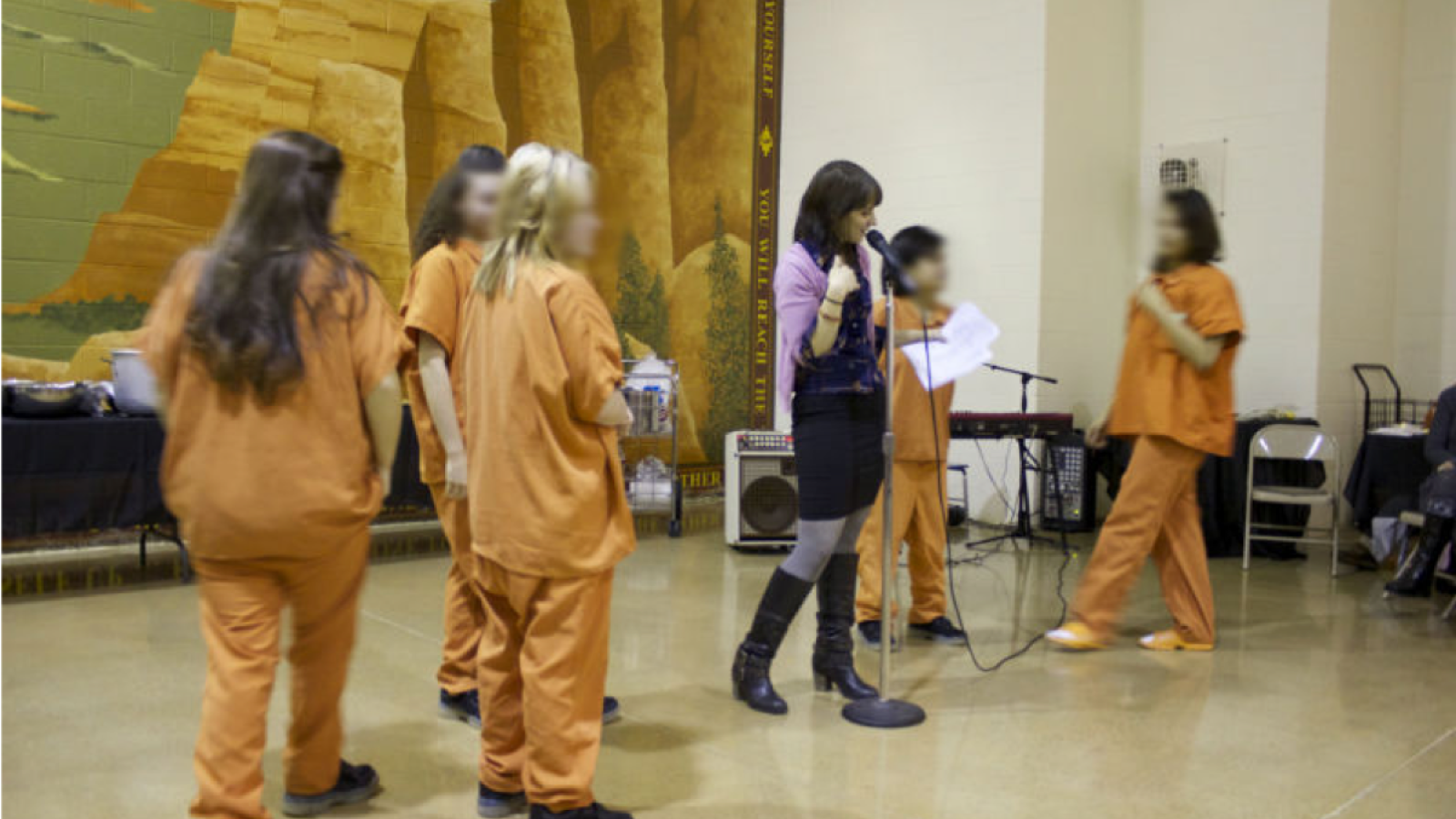 A woman with brown hair stands at the mic while girls in orange jumpsuits stand next to her.
