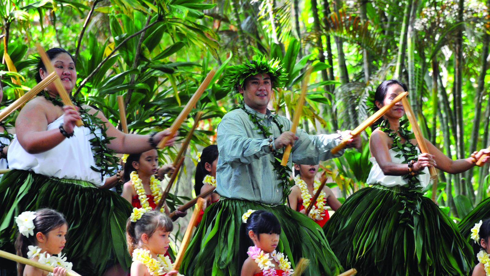 Dancers wearing grass skirts and floral wreaths holding bamboo sticks perform a traditional Hawaiian dance. 