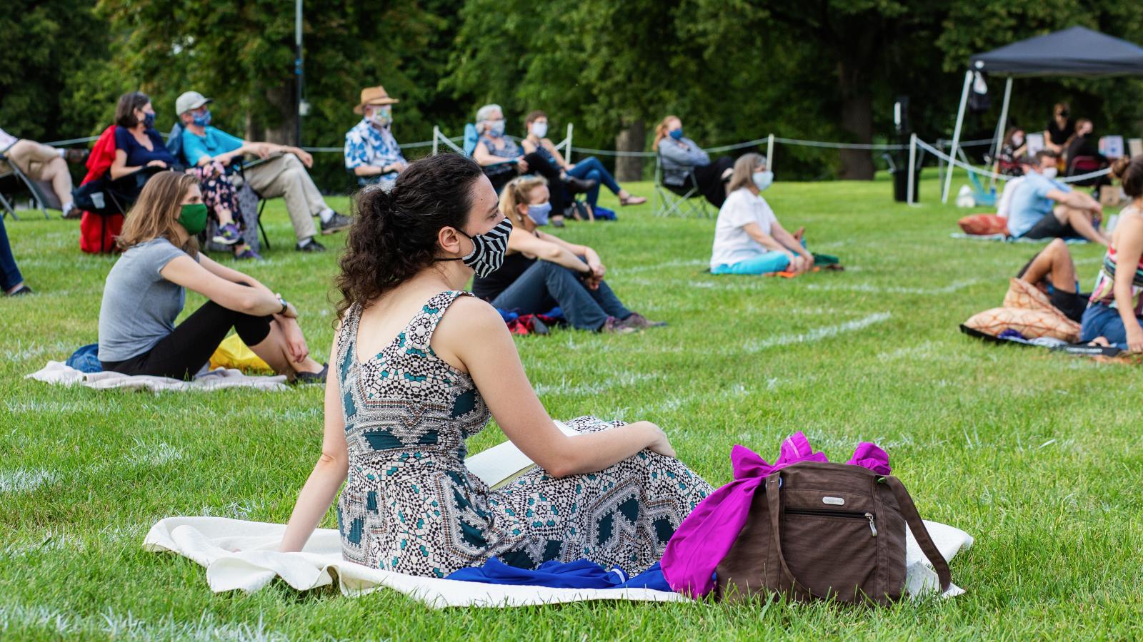 People wearing masks sit on picnic blankets on the grass while spread out from one another