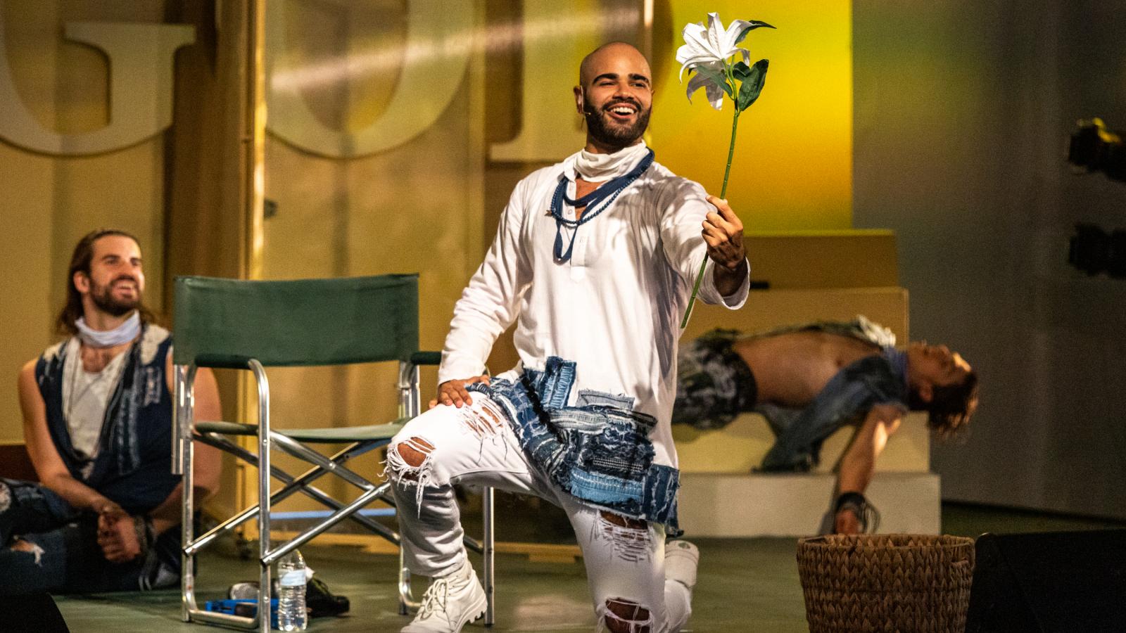 Three actors onstage, one of whom is kneeling and flourishing a flower, perform Godspell