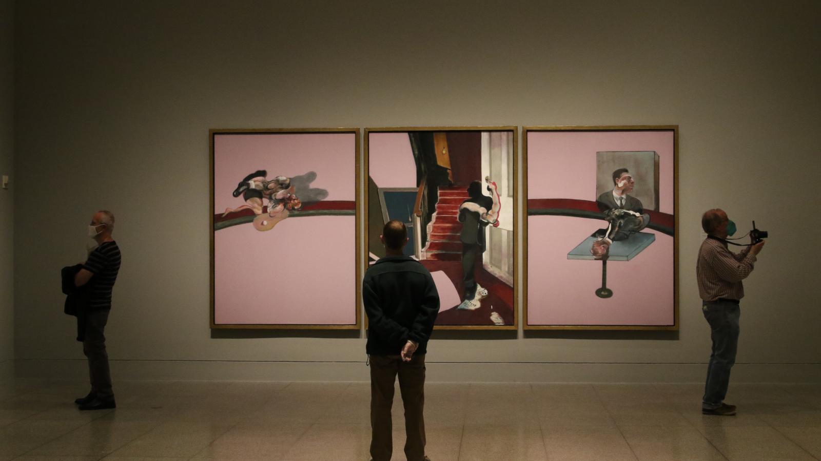 Masked museum visitors look at three large paintings by Francis Bacon