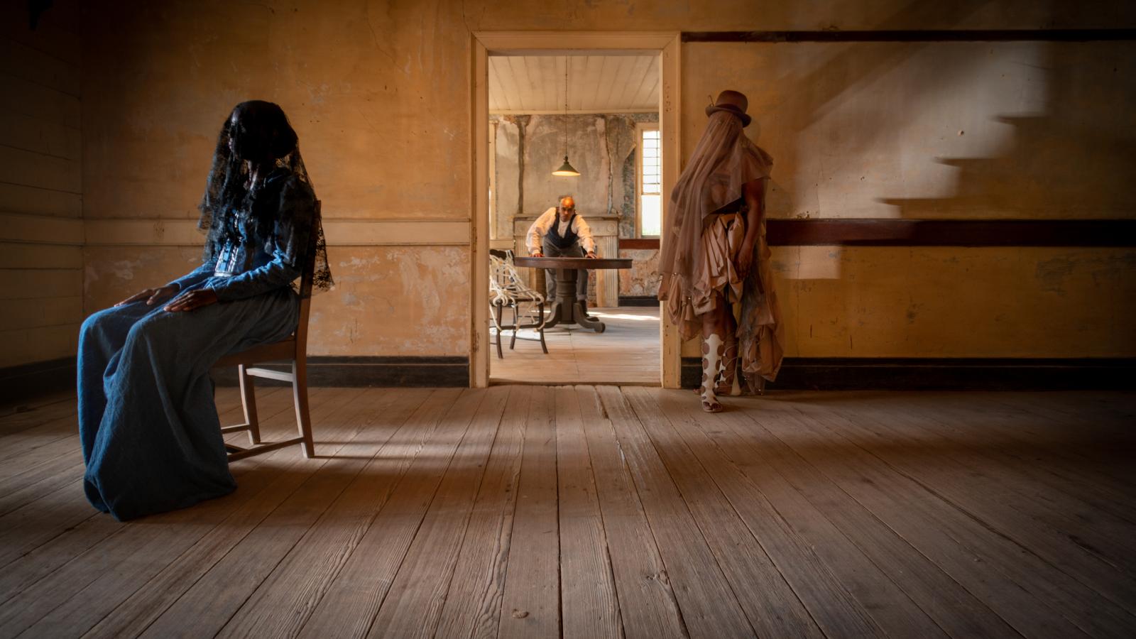 A Black woman in a blue dress and black veil sits in a chair to the left, a woman with a top hat and veil stands to the left and through the door way of a plantation mansion is a Black man standing over a table. 