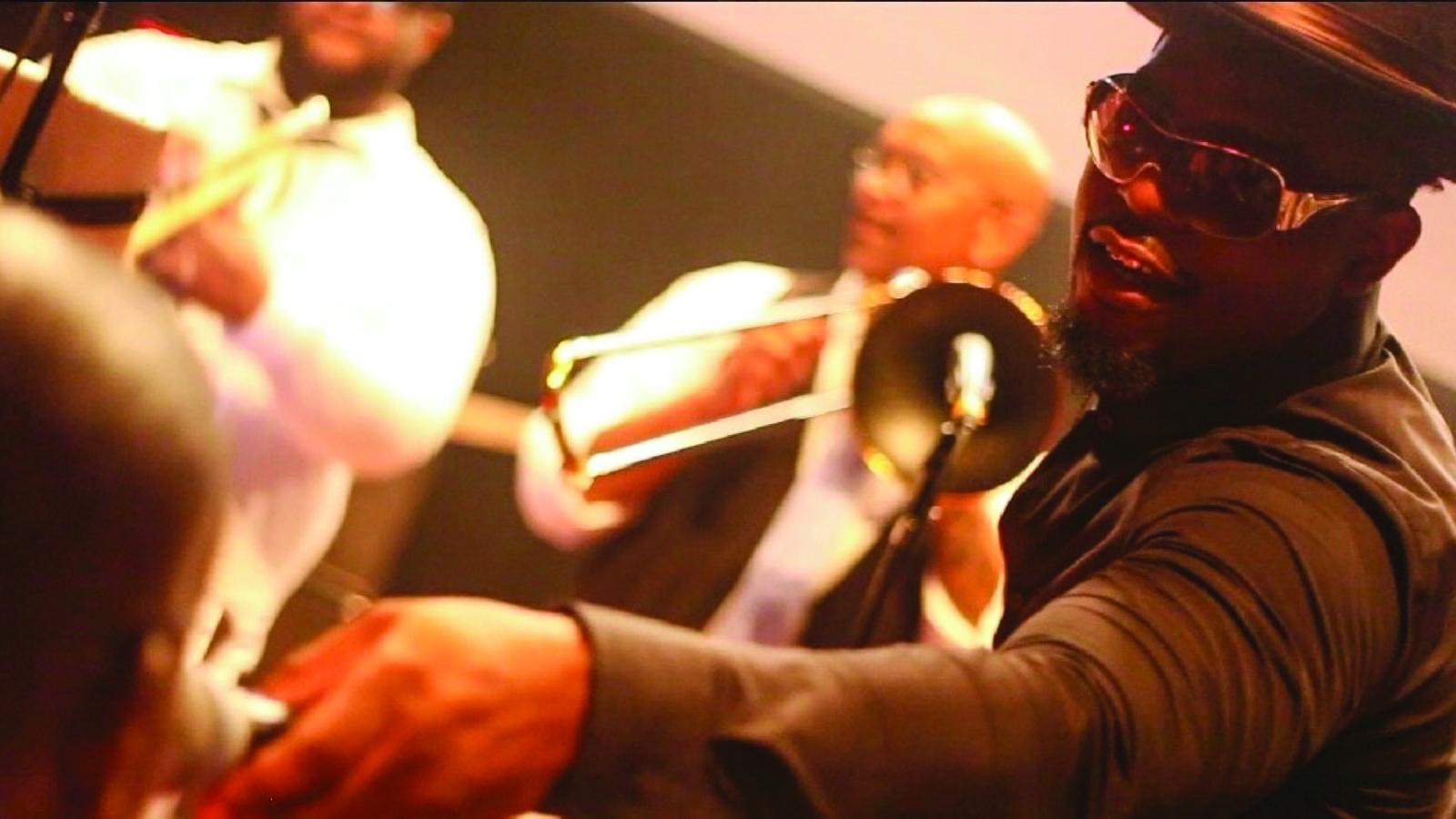 Man in sunglasses and black hat gesturing and laughing with other musicians on stage.