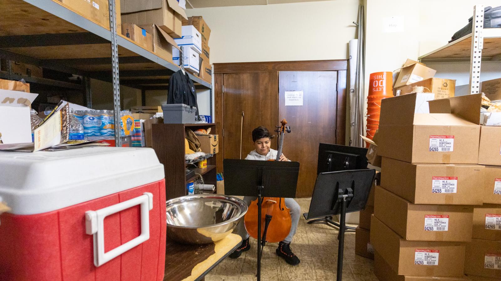 In a storage closet full of boxes and a large cooler, a boy sits behind several music stands, practicing the cello.