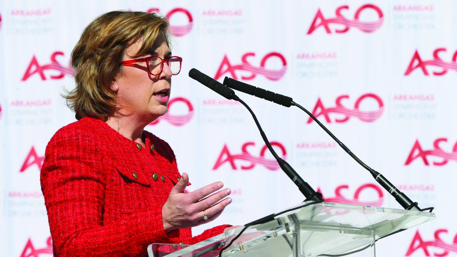 A woman speaks at a podium, a step-and-repeat banner is behind her which shows the logo for the Arkansas Symphony Orchestra.