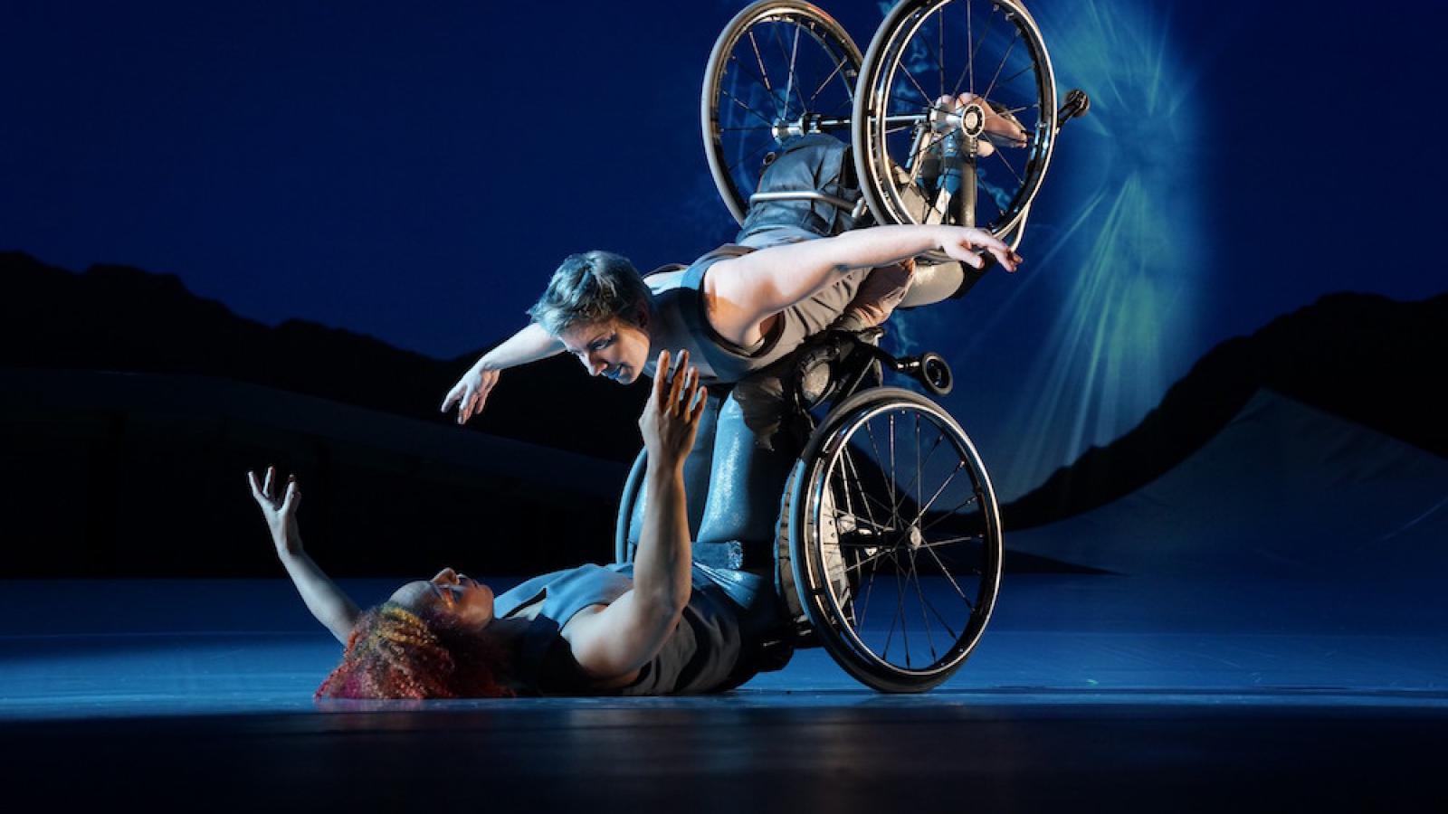 Laurel Lawson, a white woman with short cropped teal hair, is flying in the air with arms spread wide, wheels spinning, and supported by Alice Sheppard. Alice, a multiracial Black woman with coffee-colored hair, is lifting from the ground below. They are making eye contact and smiling. A burst of white light appears in a dark blue sky. 