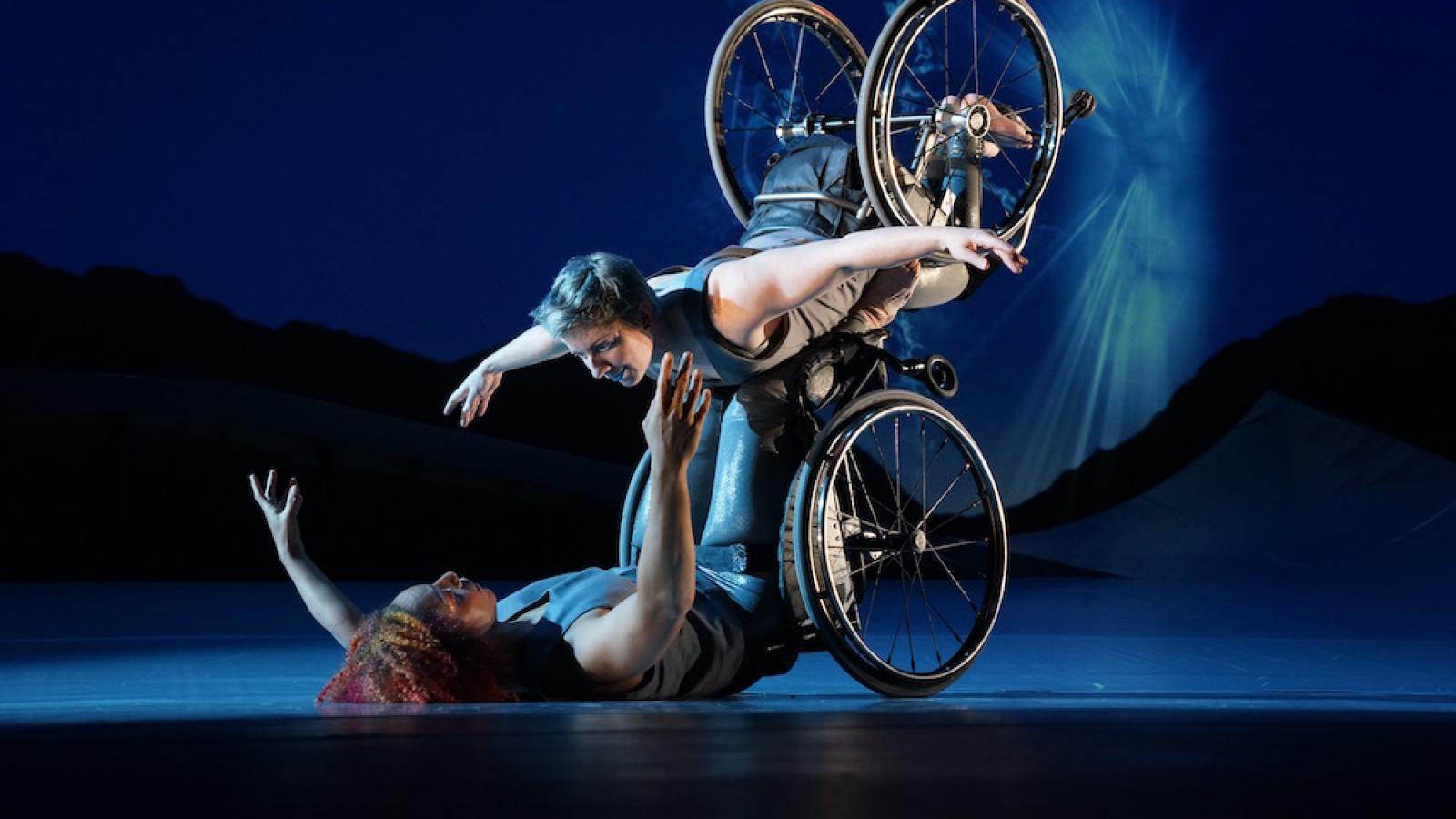 Laurel Lawson, a white woman with short cropped teal hair, is flying in the air with arms spread wide, wheels spinning, and supported by Alice Sheppard. Alice, a multiracial Black woman with coffee-colored hair, is lifting from the ground below. They are making eye contact and smiling. A burst of white light appears in a dark blue sky.