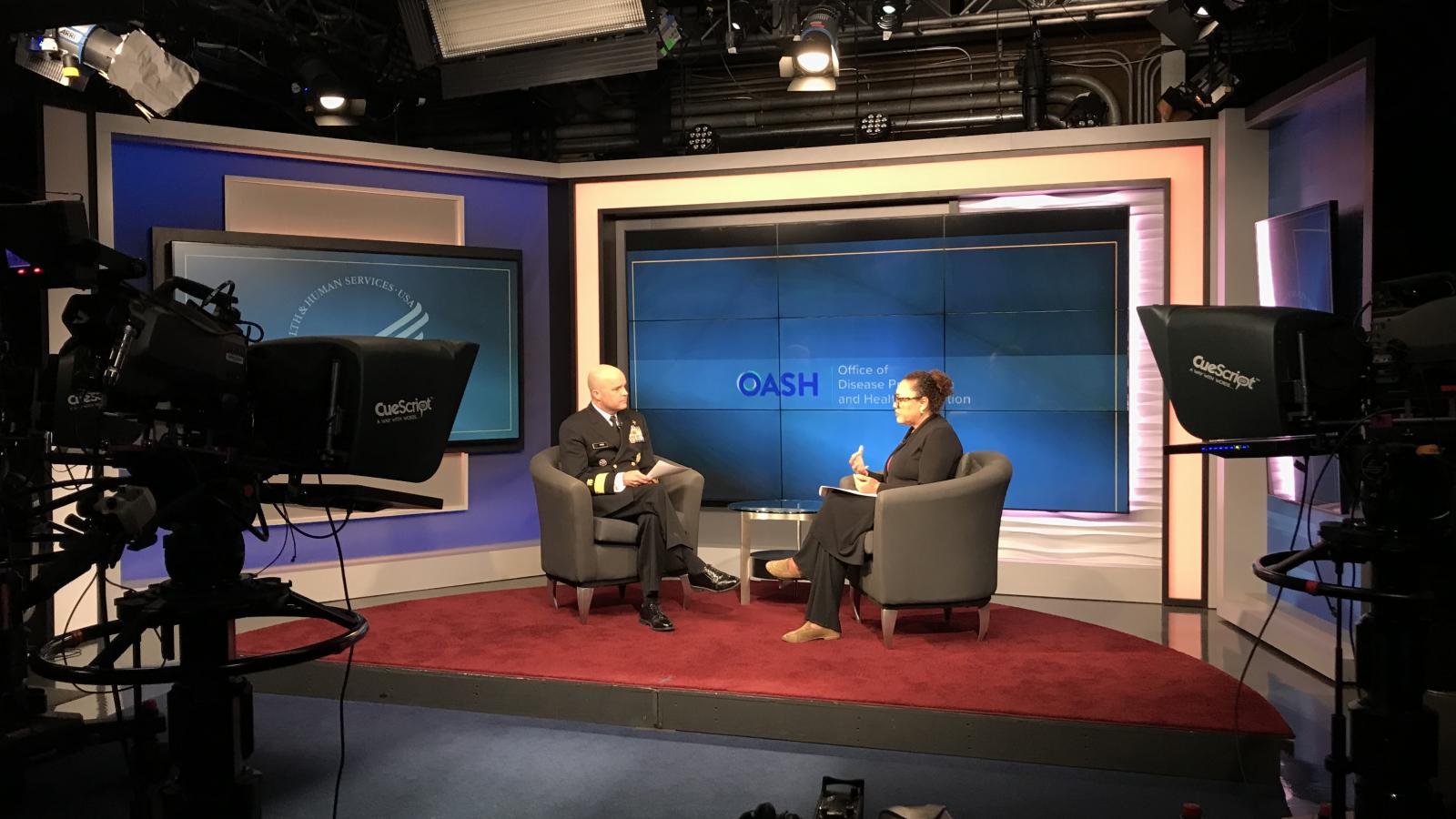 A man and woman are seated across from each other in a studio being filmed.