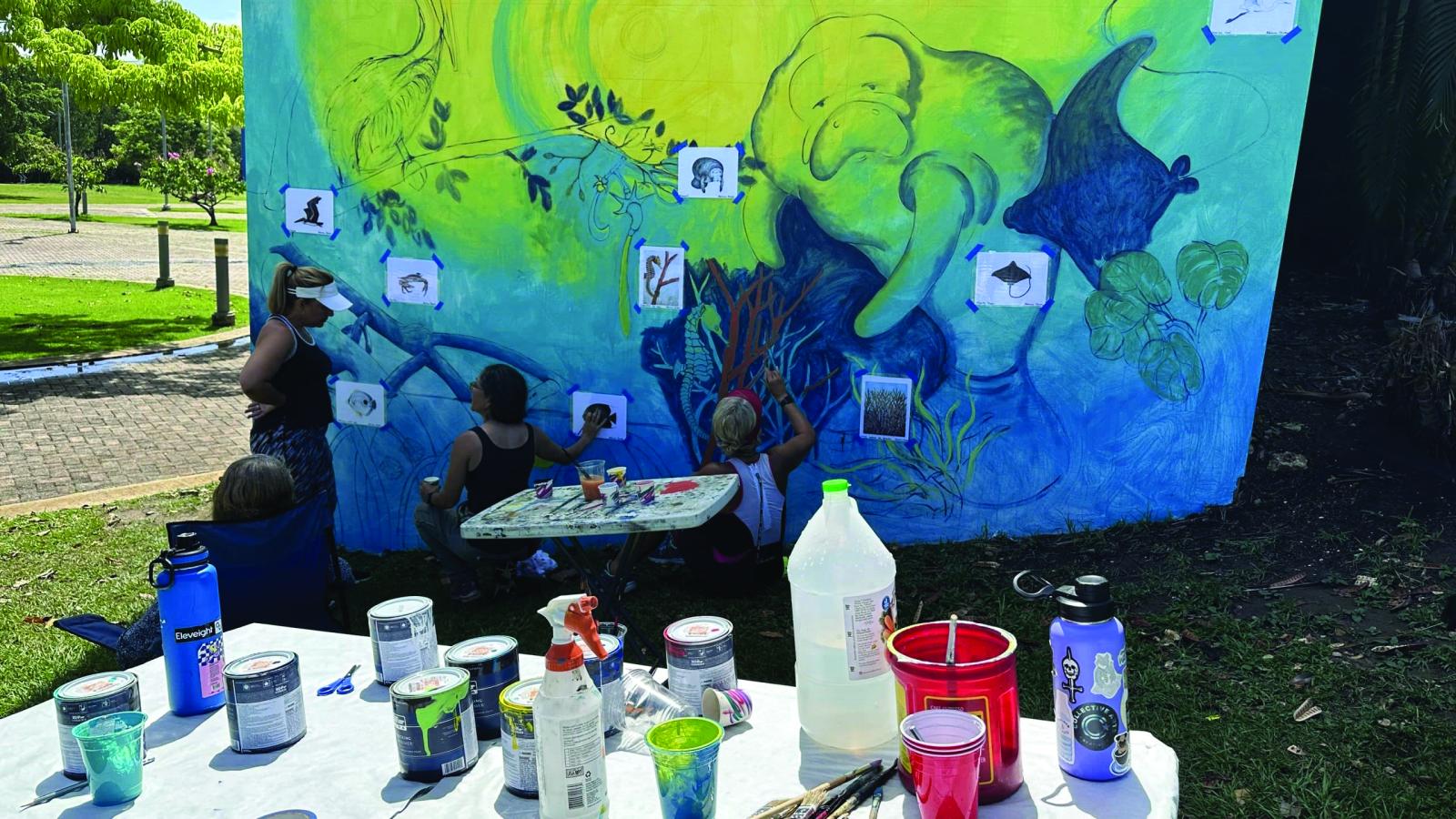 People working on an outdoor mural showing sea life. In the foreground is a table with paints and other supplies.
