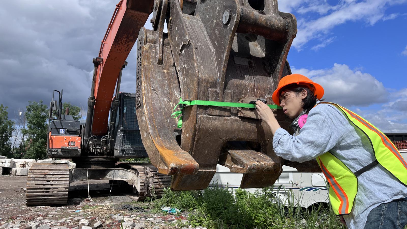 A person wearing an orange hard hat and a yellow safety vest attaches a camera to the claw of a large construction excavator. The claw has a green ribbon around it, and the person is leaning forward, peering through the camera's viewfinder.
