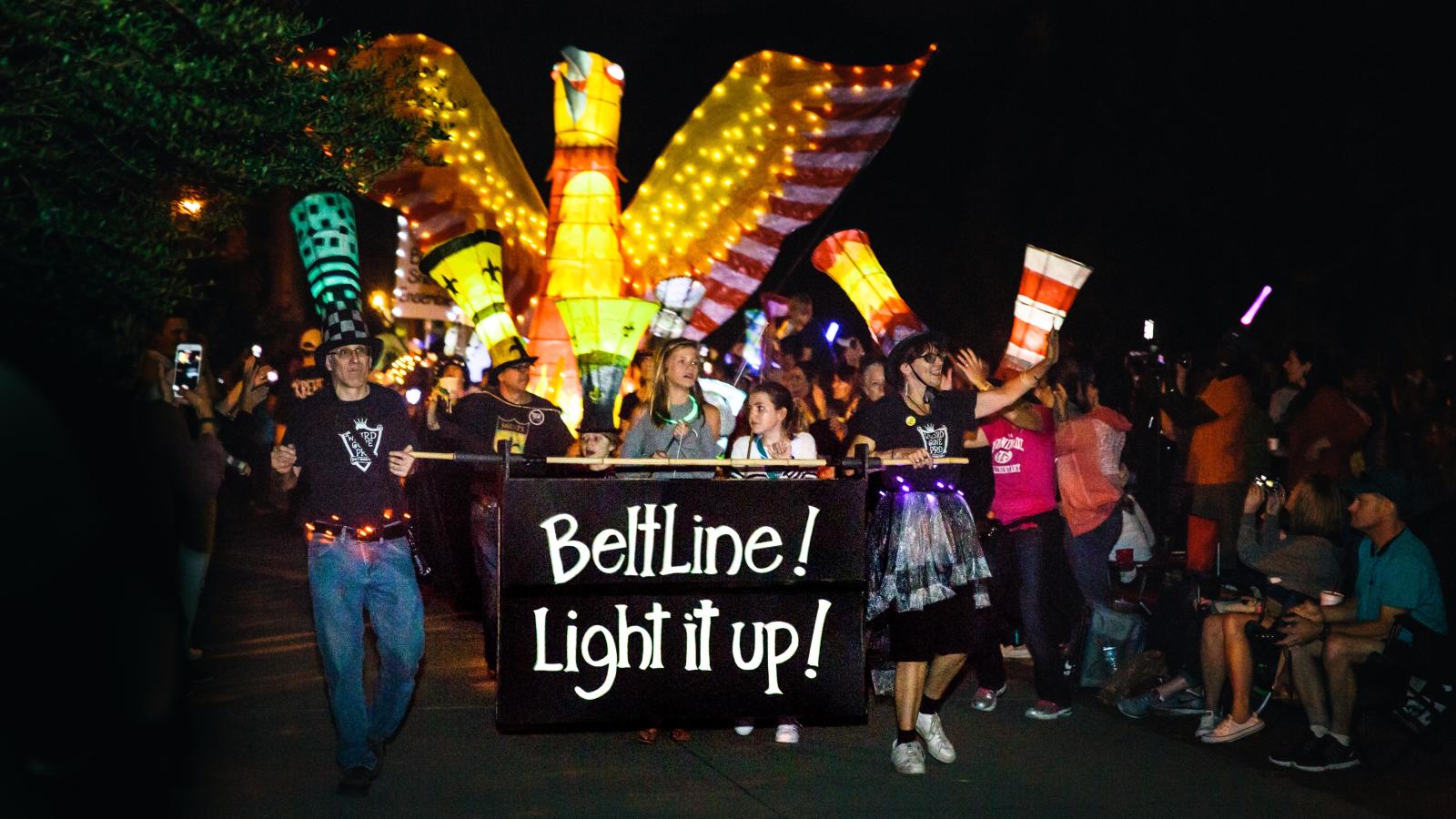 People lead a parade at night, holding a large sign that says "Beltline! Light it up!" They, as well as several others, are wearing tall hats that look like illuminated paper lanterns. Behind them is a large illuminated paper lantern of an orange and yellow bird.